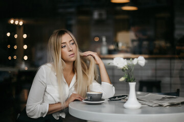 Front view of woman having coffee break while resting in the cafe. Serious woman, with blond hair, wearing formal apparel, enjoying of city view and drinking ordered coffee
