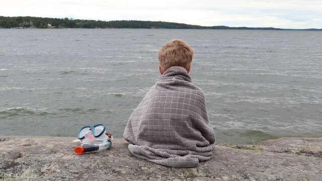 One small boy sitting on a cliff next to a lake. Summer day, but cloudy and cold. Gray towel and back towards camera. Mälaren, Stockholm, Sweden, Scandinavia, Europe.