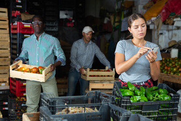Portrait of young woman working in vegetable warehouse, using smartphone to count quantity of stacks.