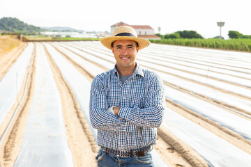 Confident smiling farmer standing against background of farm plantation mulched with polyethylene...