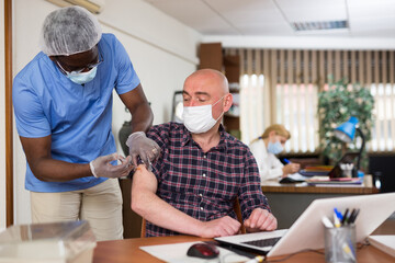 Obraz premium Medical worker who comes to the office of a large company during a pandemic vaccinates a man employee in a protective ..mask against the coronavirus