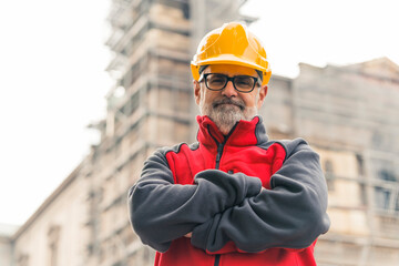 Middle-aged male architect at construction site wearing eyeglasses and hard hat looking into camera with arms crossed. Horizontal outdoor shot. High quality photo