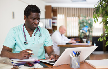 Focused african american doctor therapist works at a hospital computer, studying outpatient cards patients in the ..resident's office