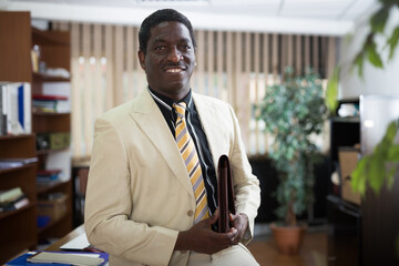 Portrait of successful confident african american businessman standing with briefcase with documents in modern office interior, looking at camera with slight smile
