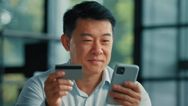 Happy Asian man client buyer businessman use banking mobile phone app at office hold plastic credit card enter secure code buy goods in internet store shopping online cryptocurrency money transaction 