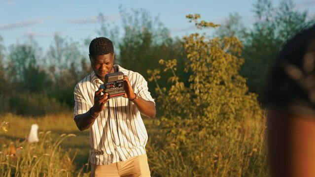 Young adult black man in field taking pictue of his friends with vintage instant film camera. Group camping. Horizontal outdoor video. High quality 4k footage