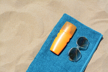 Soft blue beach towel with bottle of sunblock and sunglasses on sand, flat lay. Space for text