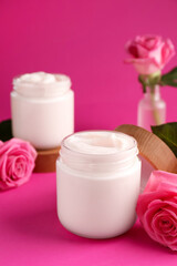 Glass jars of face cream and roses on pink background