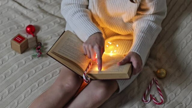 Caucasian teen girl in white knitted dress reads a book holding a finger with a burning garland across the page while sitting on a bed with a box of advent calendar and candy lollipops in the bedroom,