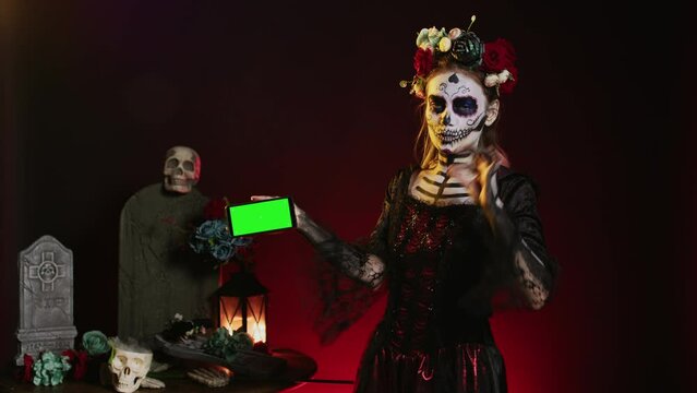 Young woman holding mobile phone with greenscreen display in studio, showing blank copyspace template. Model dressed as holy goddess with isolated chroma key mockup on smartphone.