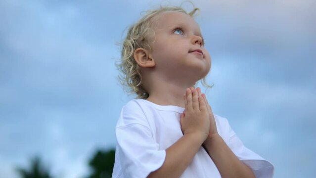 Bottom view of a little blond girl who looks like an angel. She holds her hands in front of her chest in prayer to her god. Her eyes look up to the sky.