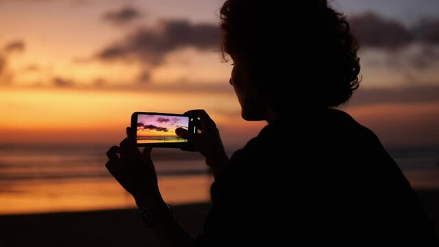 Silhouette of a woman blogger from the back against the backdrop of a sunset. A woman holds a smartphone with both hands and shoots the onset of twilight.