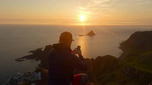 Men taking a picture with his phone of the sunset at the ocean and island Bleiksoya rock landscape in Norway