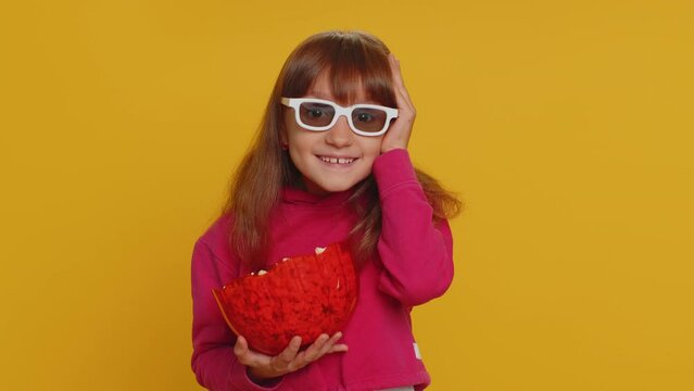 Excited young toddler school girl in 3D glasses eating popcorn, watching interesting tv serial, sport game, film, online social media movie content. Teen female child kid on studio yellow background