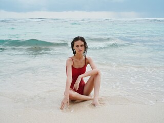 Woman with wet hair after swimming in the ocean in a red swimsuit sits on the beach in Bali and looks into the camera, a trip to Indonesia 