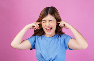 Portrait of angry irritated young woman wearing casual t-shirt covering her ears with hands and shouting while standing isolated over pink background