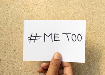 Man holding paper with text #METOO handwritten on a white card with a cork board background