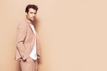 Portrait of handsome confident stylish hipster lambersexual model. Sexy modern man dressed in elegant suit. Fashion male with curly hairstyle posing in studio. Isolated on beige