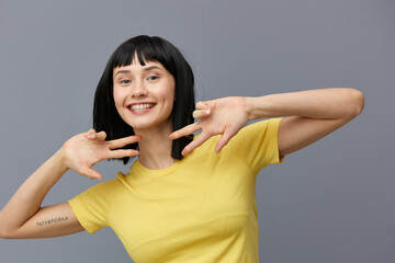Fototapeta premium a happy, sweet woman stands smiling at the camera on a gray background in a yellow T-shirt and shows a victory sign with her fingers holding her hands near her face