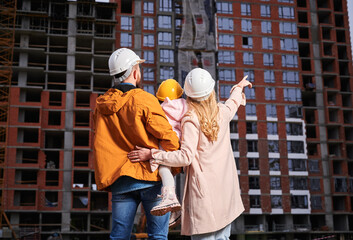 Plakat Back view of woman pointing at apartment building under constructing while standing next to husband and daughter on the street. Happy family looking at new home at construction site.