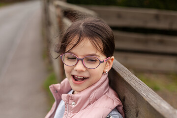 Little beautiful cute girl in glasses smiling leans on a wooden fence. Village child in a vest on a walk in the countryside