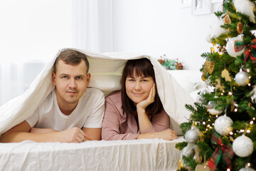 Obraz na płótnie Canvas happy couple in love lying under blanket in decorated bedroom with christmas tree
