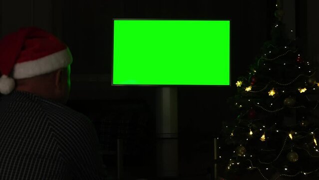 Man in Santa hat watching green screen TV mockup on black background with flashing lights of garland on Christmas. Chroma key monitor of digital device on dark backdrop on holidays in decorated room