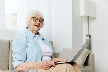 a joyful, funny old lady is sitting on a cozy sofa in a bright room and holding a laptop on her lap, working remotely despite her age