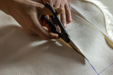 Hands of experienced worker in the handmade industry, performing cutting tasks on white fabric, on a white surface, the beginning of red hat production. Homemade sewing. Working tailor. Christmas hat.