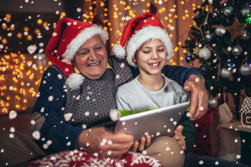 Grandfather and grandson sitting on chair and looking at digital tablet during christmas. Senior man with a kid having a video call on digital tablet.