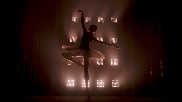 Young ballerina in white tutu dancing in pointe shoes on tiptoes against backdrop of smoke and spotlights with soft white light. Silhouette of flexible woman ballet dancer performing dance pas in dark