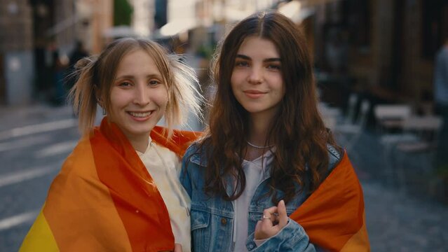 Portrait of a lesbian couple with an lgbt flag looking at the camera. Two happy female friends posing in the city centre with a rainbow flag.Woman relationships, lgbt concept