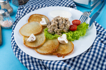 Deruny - potato pancakes with mushrooms, champignons, sour cream on a white plate. Cherry tomatoes, salad. Restaurant serving on a blue background, checkered napkin, fork, knife. Top view, copy space