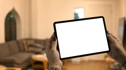 Hands holding tablet with blurred living room lighted with soft lights in background. Blank screen for your advertise design