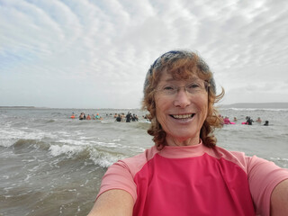 Mature woman taking a selfie before swimming in the sea. Cold or open water swimming is beneficial for both physical and mental health. She is smiling and wearing pink rash vest. Copy space on left. 