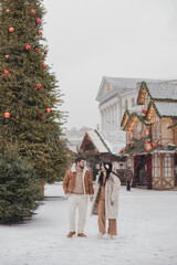 Cheerful loving couple in warm cozy clothes walk outdoors on festive city streets. Man and woman skating on ice rink as snow falls. Cold happy winter day. Holidays, Christmas, New Year, love concept.