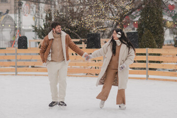 Cheerful loving couple in warm cozy clothes walk outdoors on festive city streets. Man and woman skating on ice rink as snow falls. Cold happy winter day. Holidays, Christmas, New Year, love concept.
