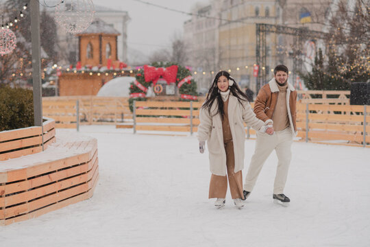 Cheerful loving couple in warm cozy clothes walk outdoors on festive city streets. Man and woman skating on ice rink as snow falls. Cold happy winter day. Holidays, Christmas, New Year, love concept.