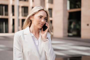 Calm blonde caucasian young woman in white suit talking by phone outdoors looking aside over blurry building on background. Attractive Italian businesswoman calling to husband during break. Business