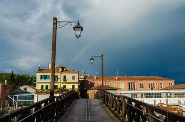Fototapeta na wymiar L'arcobaleno attraversa il cielo di Venezia dopo la pioggia su un ponte di legno con dei lampioni 