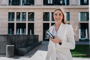 Pretty blonde lawyer woman going to work holding diary and phone dressed in white suit. Cheerful attractive Italian businesswoman grateful for her career. Successful European female waling outside.