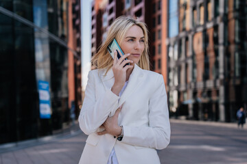 Confident Italian businesswoman in white suit talking by phone outside against blurry huge buildings. Attractive young lawyer making call to client. Successful blonde European female walking