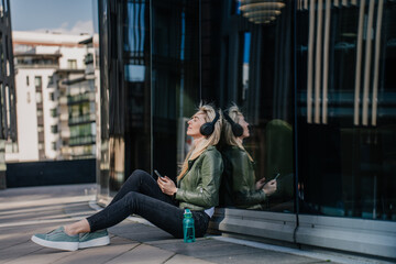 Fototapeta premium Cheerful young blonde woman in headphones wearing casual clothes sitting on the ground with water bottle relaxing listening music on weekend. Cheerful italian hipster girl enjoying life and youth.