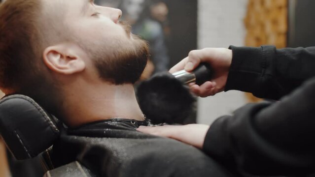 Master works with a brush beard of men in chair the barbershop, hairdresser makes beardstyle for a young man indoor. After a professional barber care clients