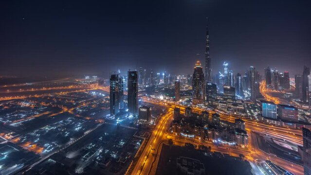 Aerial panorama of tallest towers in Dubai Downtown skyline and highway night timelapse with rising Moon. Financial district and business area in smart urban city. Skyscraper and high-rise buildings