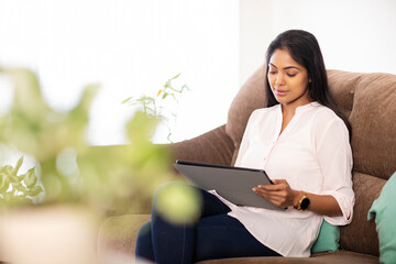 Fototapeta premium Pretty Indian young woman using digital tablet sitting on sofa at home.
