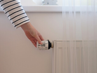 Woman regulates temperature at home with the heating thermostat to save energy, close-up with hand. Temperature control valve on radiator. White heater on wall with curtain in modern apartment.