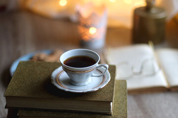 Plate of chocolate chip cookies, stack of vintage books, reading glasses, cup of tea or coffee, lit candle and fairy lights. Hygge at home. Selective focus.