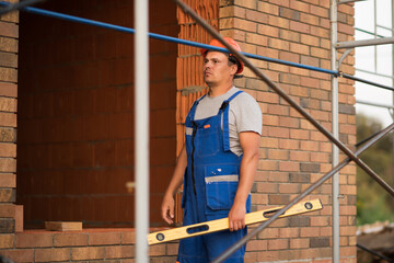 a male builder in blue overalls and a construction helmet stands near a window opening with a level, building a house