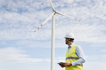 Caucasian engineer holding digital tablet and turning towards a wind turbine